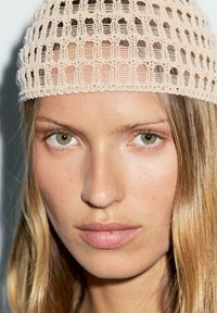 Cappello a maglia beige con un design a maglia aperta poggia su lunghi capelli ondulati, incorniciando un viso dai toni neutri con occhi verde chiaro.