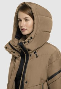 Gaisa pūkains jaka gaiši brūnā krāsā, ar lielu kapuci, ar rāvējslēdzēju un melniem akcentiem, kā arī ar pogām. Tekstūras audums ar quilted dizainu.