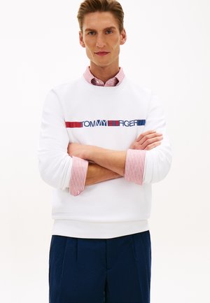 Mann mit verschränkten Armen, der einen weißen Sweatshirt mit rotem und blauem Tommy Hilfiger-Logo, rot gestreifte Hemdärmel und marineblaune Hose trägt.