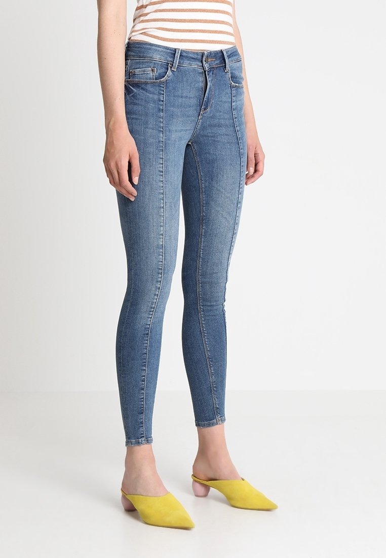 Jean skinny en denim bleu moyen avec une texture lisse, taille haute et décoloration subtile ; porté avec des mules jaunes à enfiler.