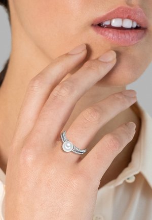 Silberner Ring mit einer Perle in der Mitte, bestehend aus zwei verflochtenen Bändern, die mit kleinen, klaren Steinen verziert sind, auf einer Hand präsentiert.