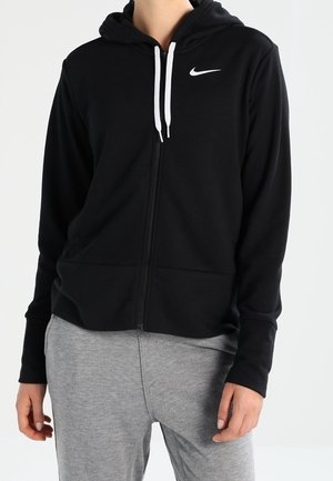 Person iført en sort Nike hættetrøje med lynlås med hvide snore og lysegrå joggingbukser, stående mod en ensfarvet baggrund.