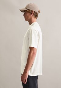 Biały T-shirt z krótkim rękawem wykonany z miękkiej tkaniny. Posiada okrągły dekolt, luźny krój oraz boczne rozcięcie. Noszony z beżową czapką i ciemnymi spodniami.