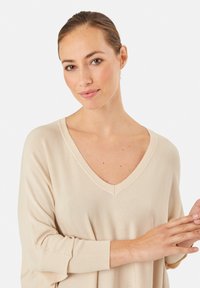 Maglione beige con scollo a V, maniche larghe e polsini a coste. Tessuto morbido e vestibilità rilassata. Il modello indossa il capo con le braccia visibili.