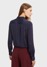 Camicia blu navy con strisce verticali bordeaux, maniche lunghe e colletto. Dotata di polsini con bottoni e un design con yoke sul retro. Tessuto dalla texture liscia.