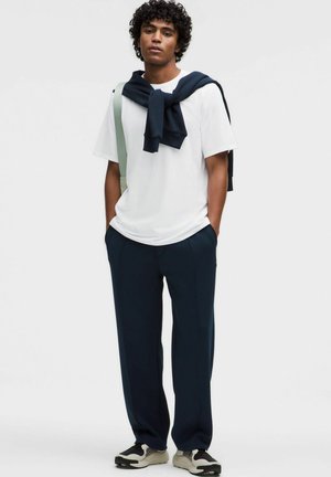 Valkoinen lyhythihainen t-paita, laivastonsiniset housut, kaulassa solmittu laivastonsininen neule, jalassa mustavalkoiset lenkkarit, puhtaata taustaa vasten poseeraamassa.