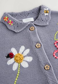 Cardigan gris en tricot avec des boutons en bois en forme de fleur, marguerite blanche brodée avec coccinelle, abeille et motifs floraux colorés sur le col.