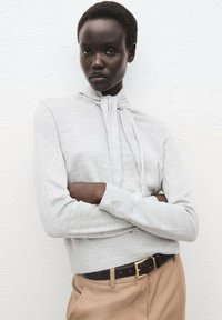 Maglione attillato grigio chiaro con collo alto e maniche lunghe, caratterizzato da un dettaglio legato al collo, abbinato a pantaloni beige e una cintura marrone.