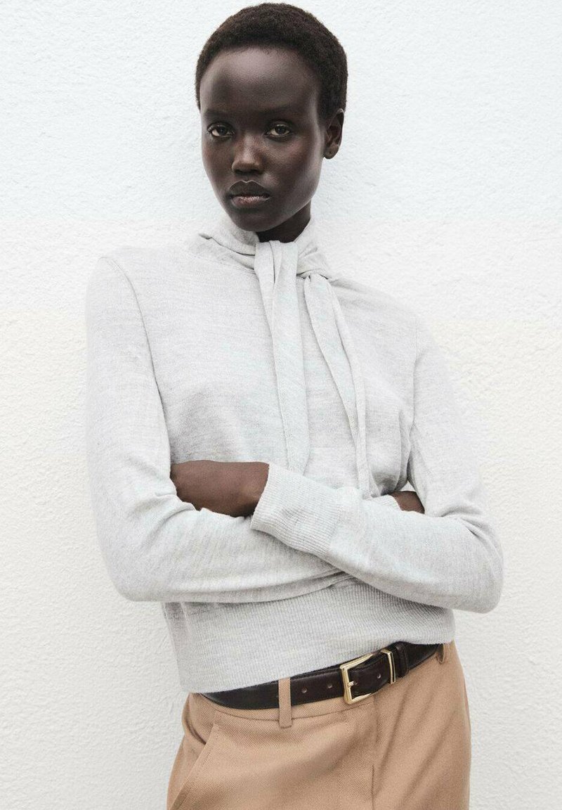 Hellgrauer, figurbetonter Pullover mit hohem Hals und langen Ärmeln, der mit einem gebundenen Detail am Kragen versehen ist, kombiniert mit beigen Hosen und einem braunen Gürtel.