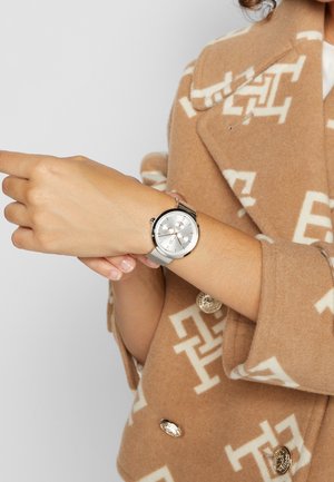 Person iført en beige mønstret frakke justerer et sølv armbåndsur med mesh-rem på venstre håndled mod en ensfarvet baggrund.