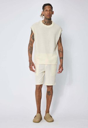 Homme avec des tatouages portant un haut en maille crème sans manches et un short assorti avec des chaussures beige slip-on, debout sur un fond blanc.