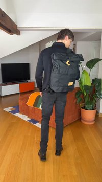 Mann iført svarte klær bærer en stor svart ryggsekk i stuen med oransje sofa, potteplante og TV.