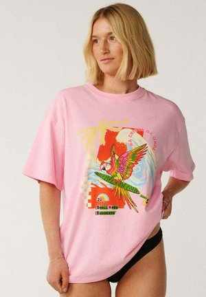 Donna bionda che indossa una T-shirt rosa oversize con stampa colorata di pappagallo e motivo tropicale, abbinata a slip bikini neri.