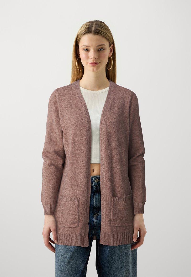 Gilet rose avec un col en V, des manches longues, des poignets côtelés et deux poches avant. Porté par-dessus un top blanc et associé à un jean taille haute.