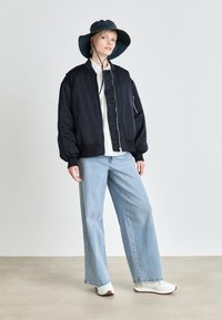 Schwarze Bomberjacke mit gerippten Bündchen, kombiniert mit hellblauen weit geschnittenen Jeans und weißen Sneakers. Das Model trägt einen dunkelgrünen Bucket Hat.