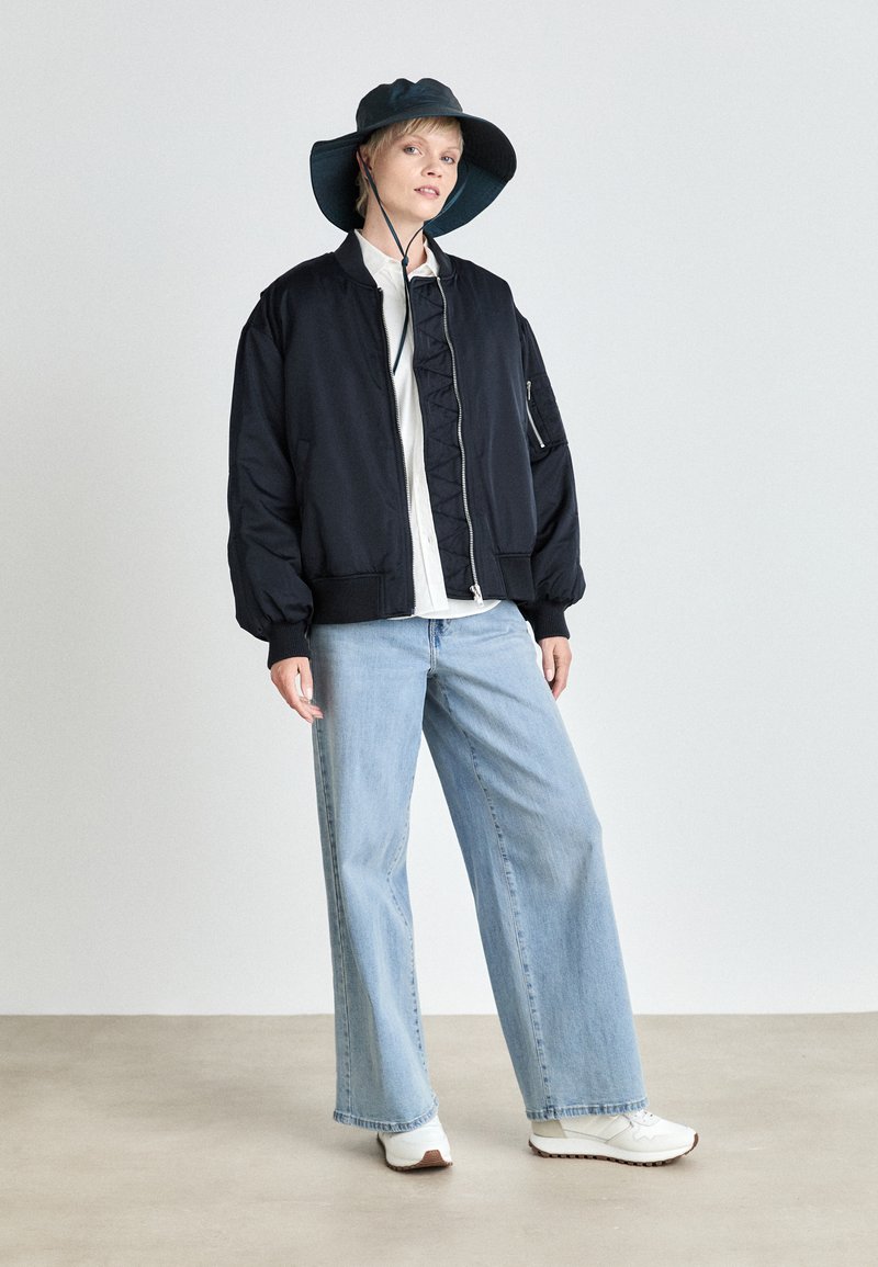 Schwarze Bomberjacke mit gerippten Bündchen, kombiniert mit hellblauen weit geschnittenen Jeans und weißen Sneakers. Das Model trägt einen dunkelgrünen Bucket Hat.
