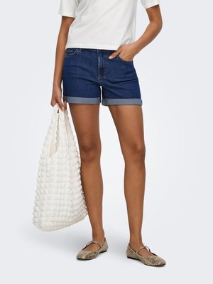 Personne portant un short en jean bleu foncé retroussé, un haut blanc et des ballerines à motif peau de serpent, tenant un grand sac blanc texturé.