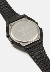 Crna nehrđajući čelik sat s teksturiranom narukvicom, s kružnim kućištem i ugraviranim adidas logom na poliranom metalnom stražnjem dijelu.