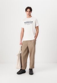 Λευκό T-shirt με μπλε γραμματοσειρά, μπεζ φαρδιές παντελόνες και μαύρα αθλητικά παπούτσια. Κρατάει μια καμβά τσάντα. Απλό φόντο.