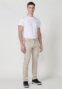 Jeans slim fit beige con una textura suave y un ligero desvanecimiento. Combinados con una camiseta blanca ajustada y zapatillas negras. Diseño y colores simples.