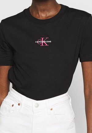 Black cotton T-shirt featuring a front pink embroidered logo "CALVIN KLEIN JEANS" with accents. Short sleeves and round neckline. White jeans.