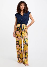 Femme debout, une main dans la poche, portant une chemise sans manches nouée bleu marine et un pantalon floral ample taille haute avec des sandales.