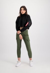 Svart cropped fleece-jakke med glidelås og sidelomme. Olivengrønne cargo-bukser med lommer og sømdetaljer. Hvite sneakers. Enkel, nøytral bakgrunn.