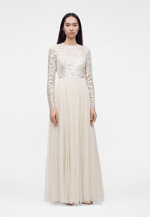 Une femme se tient debout portant une longue robe crème avec un corsage transparent brodé de sequins, des manches longues et une jupe plissée à motif à pois.