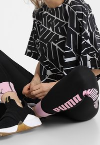 En person sitter i korslagda ben och bär svarta och rosa Puma leggings, en svart och vit mönstrad skjorta samt svarta slip-on sneakers med gummikapslar.