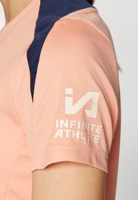 T-shirt de sport rose clair en tissu texturé, avec un empiècement bleu marine sur l'épaule et le logo "INFINITE ATHLETE" en blanc sur la manche.