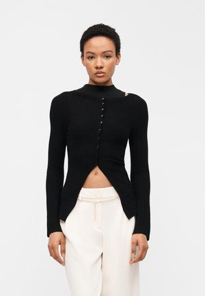 Woman wearing a fitted black buttoned long-sleeve top over a black undershirt with white pants and a thin chain belt, standing against a plain background.