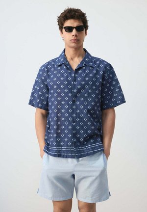 Hombre con gafas de sol negras, camisa azul de manga corta con botones y patrón, y pantalones cortos azul claro, de pie con las manos en los bolsillos.