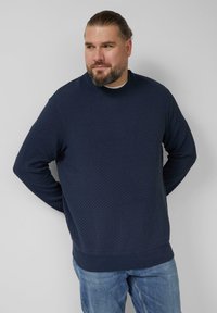 Granatowa bluza z teksturowanym wzorem diamentowym, z prążkowanym, okrągłym dekoltem i ściągaczami na rękawach i dole. Nosi się ją na jasnych dżinsach.