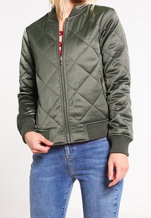 Kobieta ubrana w zieloną pikowaną kurtkę typu bomber na czerwonym topie i niebieskich dżinsach, stojąca z jedną ręką przy zamku kurtki.