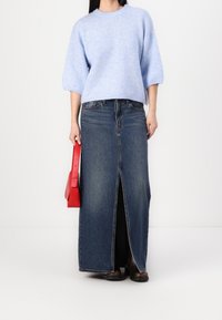 Pull en maille bleu clair à manches courtes, associé à une longue jupe en denim avec une fente à l'avant, et accessoirisé d'un sac à main rouge.