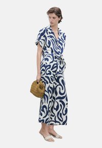 Navy and white patterned jumpsuit in lightweight fabric featuring short sleeves, a tie waist, and wide-leg silhouette. Model holds a woven bag.