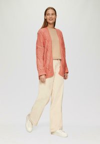 Korallfarbener Strickcardigan mit offener Vorderseite, lässiger Passform und strukturiertem Muster. Getragen über einem beigen Oberteil und kombiniert mit hellbeigen weiten Hosen und weißen Sneakern.