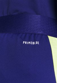 Zbliżenie na ciemnoniebieską tkaninę z teksturowaną taśmą, z białym napisem "PRIMEBLUE" i żółtym paskiem po prawej stronie.