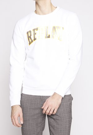 Man wearing white sweatshirt with "REPLAY" text in gold and brown, paired with gray plaid trousers, standing against a light background.