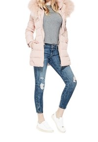 Mujer vistiendo un abrigo acolchado rosa claro con capucha de cuero, camiseta gris, jeans skinny azules desgastados y zapatillas blancas, de pie con una pierna flexionada.