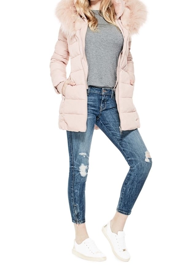 Mujer vistiendo un abrigo acolchado rosa claro con capucha de cuero, camiseta gris, jeans skinny azules desgastados y zapatillas blancas, de pie con una pierna flexionada.