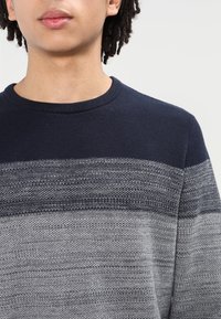 Person klädd i en marinblå och grå stickad tröja med strukturerade horisontella ränder, synlig från axlarna till läpparna mot en enkel bakgrund.