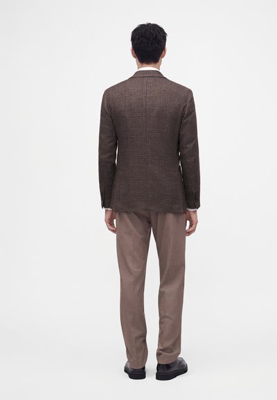 Homme debout de dos portant un blazer à carreaux marron, un pantalon marron clair et des chaussures noires sur un fond blanc uni.