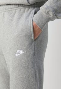 Person i ljusgrå Nike joggingbyxor med handen i fickan, med vit Nike-logotyp på vänster lår, mot en enhetlig ljus bakgrund.