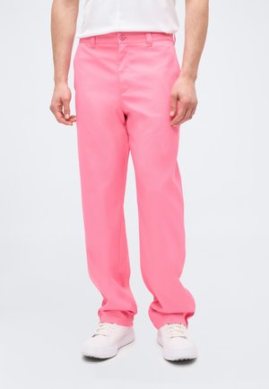 Man draagt losse roze broek, witte sneakers en een wit overhemd tegen een effen lichte achtergrond.