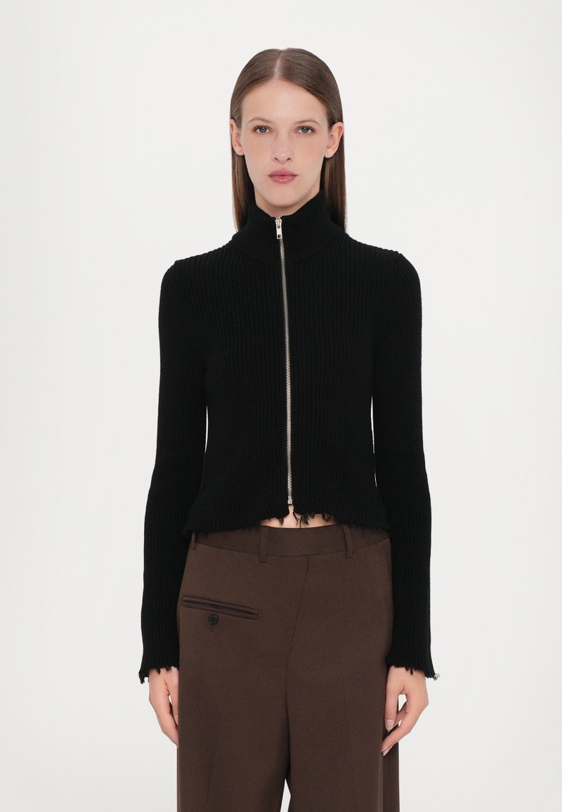 Zwarte geribde gebreide zip-up trui met een hoge kraag, cropped lengte en rafelige zoomdetails. Gecombineerd met bruine op maat gemaakte broek.