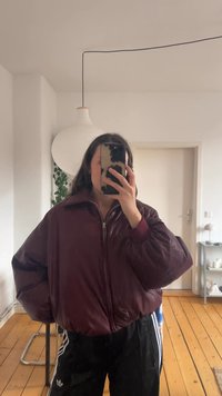 Giacca bomber bordò con chiusura a zip e colletto alto, caratterizzata da maniche gonfie e una texture lucida, indossata sopra pantaloni scuri a righe.