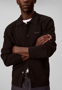 Homme bras croisés portant un cardigan noir boutonné sur une chemise blanche et un pantalon foncé sur un fond clair uni.