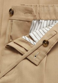 Gros plan sur la ceinture d’un pantalon beige avec bouton, fermeture éclair et doublure intérieure rayée visible.