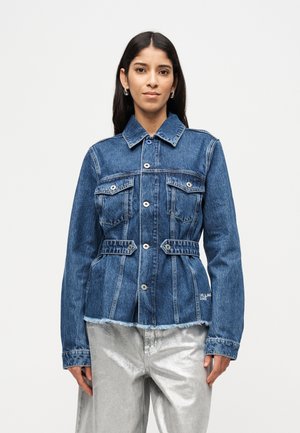 Nainen pukeutuneena istuvaan siniseen farkkutakkiin, jossa on napit, ja hopeiseen housuihin, seisoo yksinkertaisen valkoisen taustan edessä.